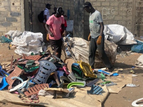 Zwei Personen aus dem Senegal sortieren bunte Kunststoffteile auf einer Plane im Freien. Im Hintergrund sind große Säcke und eine Mauer zu sehen.