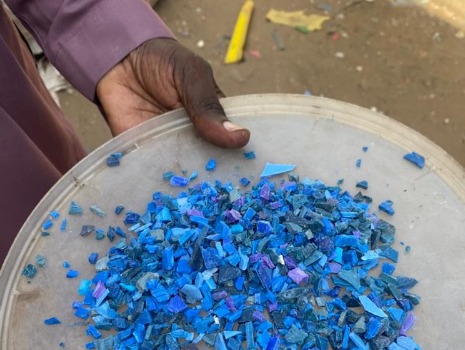 Eine Hand hält einen runden Behälter mit blauen Kunststoffgranulatstücken. Der Hintergrund ist unscharf und zeigt eine Außenfläche.