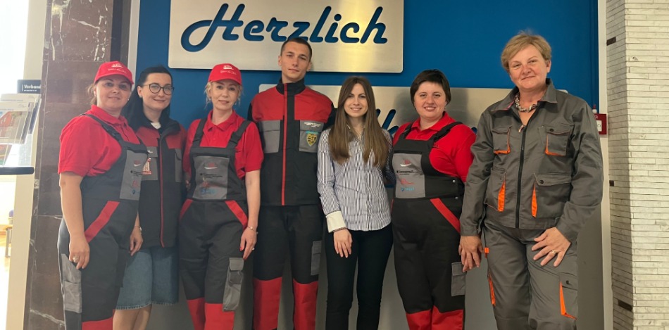 Sieben Personen, darunter Auszubildende und Ausbilder, posieren vor einer blauen Wand mit einem „Herzlich willkommen“-Schild. Einige tragen rote und graue Arbeitskleidung mit Logos, andere sind in zivil gekleidet.