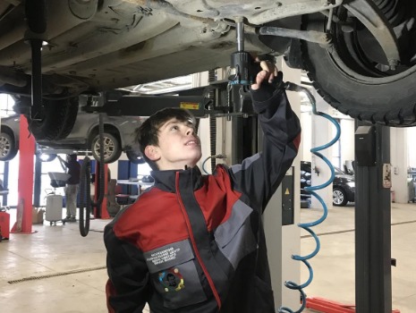 Beschreibung: Ein junger Auszubildender in unserer Arbeitsjacke benutzt ein Druckluftwerkzeug, um eine Schraube an einem hochgehobenen Fahrzeug in einer Autowerkstatt zu befestigen.