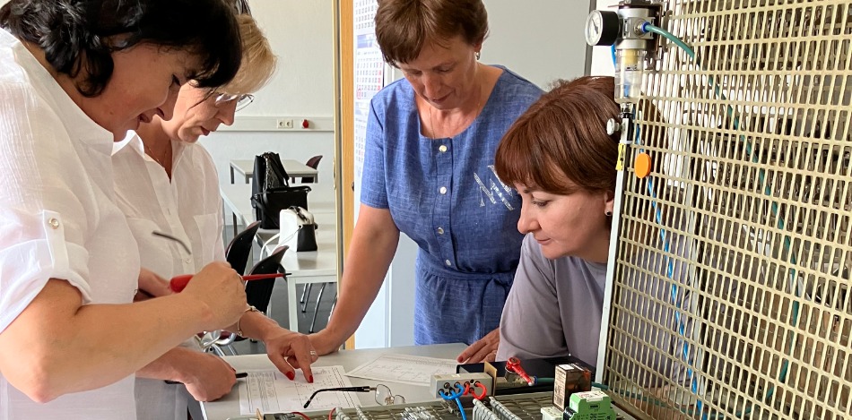 Vier Frauen arbeiten an einem großen technischen Schaltbrett mit vielen Kabeln und Bauteilen. Sie analysieren gemeinsam einen Schaltplan, während sie Werkzeuge in den Händen halten.