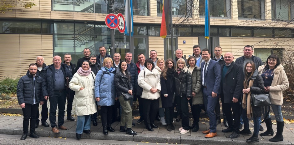 Eine Gruppe von Männern und Frauen steht vor einem modernen Gebäude mit Glasfassade. Im Hintergrund sind die deutsche und die ukrainische Flagge gehisst. Die Teilnehmer tragen warme Winterkleidung und schauen in die Kamera.