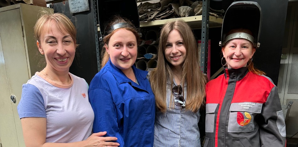 Zwei Frauen in Arbeitskleidung, darunter eine mit einem hochgeklappten Schweißerhelm, stehen mit zwei zivil gekleideten Frauen in einer Werkstatt.