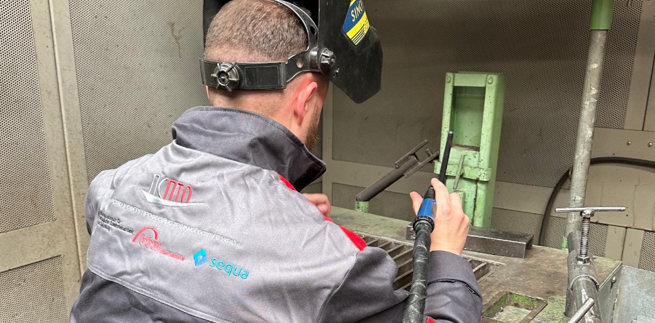 Ein Mann mit einem hochgeklappten Schweißhelm sitzt auf einem Hocker in einer Metallkabine und hält ein Schweißgerät in der Hand.
