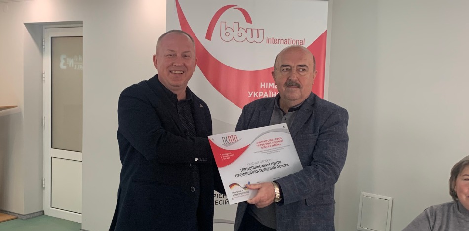 Zwei Männer schütteln sich die Hände und halten eine Plakette mit dem Logo von „bbw International“ in einem Konferenzraum.