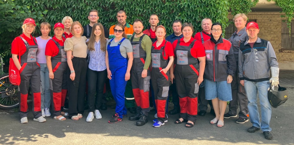 Eine Gruppe von Männern und Frauen steht vor einer grünen, mit Efeu bewachsenen Wand. Viele tragen rote und graue Arbeitskleidung mit Logos, andere sind in Freizeitkleidung. 