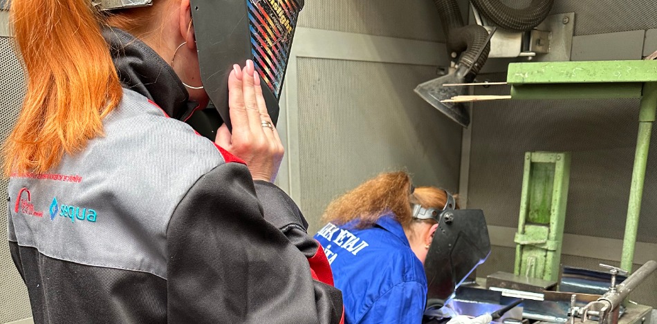 Eine Frau mit hochgeklapptem Schweißerhelm beobachtet eine andere Person, die in blauer Arbeitskleidung ein Metallstück schweißt. Die Szene findet in einer Werkstattkabine mit Belüftungssystem statt.