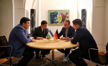 Vier Männer sitzen an einem runden Tisch und beraten. Auf dem Tisch liegen Schreibutensilien und kleine Länderflaggen (Usbekistan, Deutschland). Im Hintergrund hängt ein abstraktes Bild. Die Szene wirkt formell und sachlich.