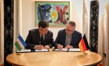 Zwei Männer sitzen an einem Tisch und unterschreiben Dokumente. Usbekische und deutsche Flaggen stehen auf dem Tisch. Hintergrund: modernes Büro, abstraktes Bild an der Wand. Geschäftsabschluss zwischen Usbekistan und Deutschland.