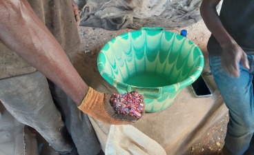 Hand mit Handschuh hält bunte Plastikstücke neben grünem Waschbecken; Umgebung wirkt wie Recycling-Werkstatt
